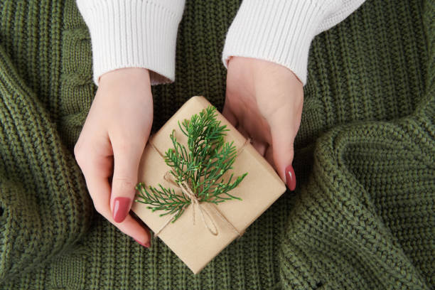 female hands holding a kraft gift box with fir decoration on knitted green sweater. 