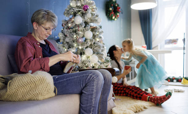 Senior woman is doing craft crocheting, making jute baskets sitting on a sofa in a living room decorated with a Christmas Tree, while her adult daughter with a little granddaughter are playingin the backdrop.