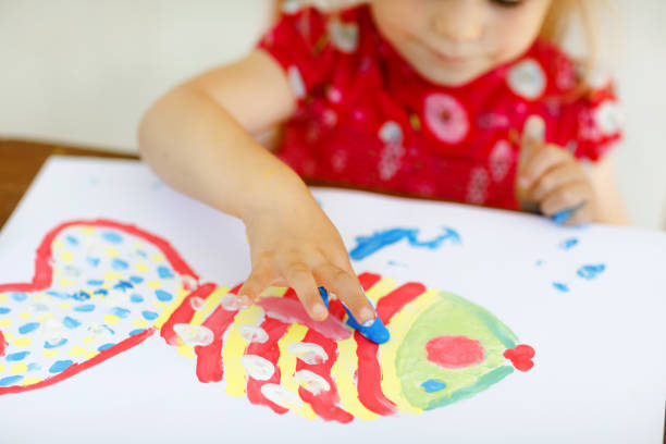Little creative toddler girl painting with finger colors a fish. Active child having fun with drawing at home, in kindergaten or preschool. Education and distance learning for kids. Zero-waste art and crafts.