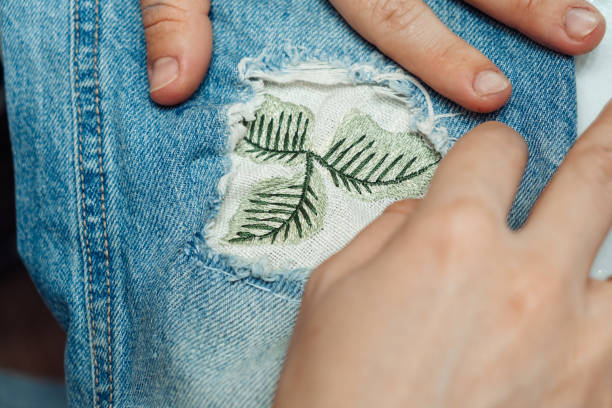 A woman mends jeans, sews a patch on a hole, hands close-up. Mending clothes concept, reusing old jeans.