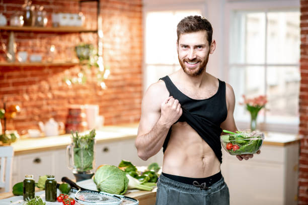 Portrait of a handsome sports man showing muscles while eating healthy vegetarian salad on the kitchen at home