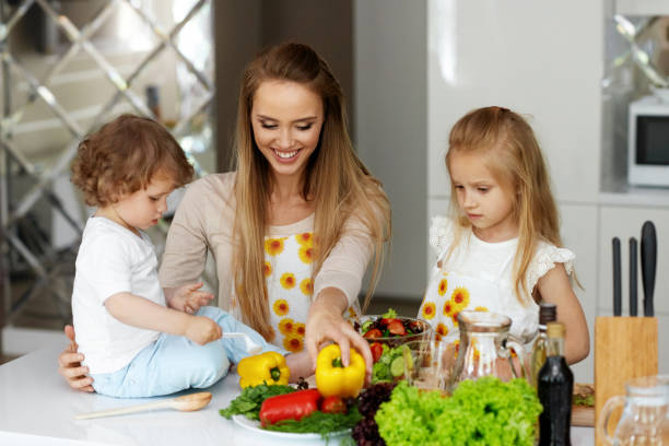 Woman With Children Cooking Healthy Food In Kitchen. Happy Smiling Mother With Daughter And Son Preparing Salad With Vegetables At Home. Family Healthy Nutrition. Vegan Diet for Children.